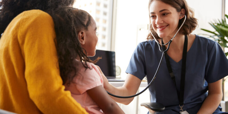 Female Pediatrician Wearing Scrubs Listening To Girls Chest With Stethoscope In Hospital Office