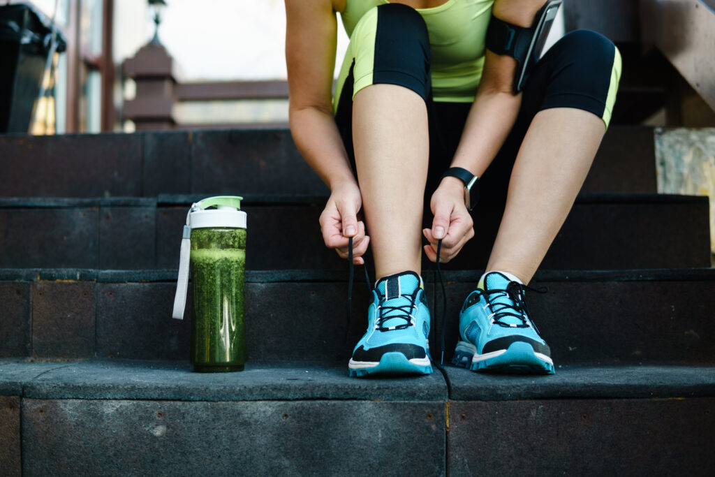 Green detox smoothie cup and woman lacing running shoes before workout