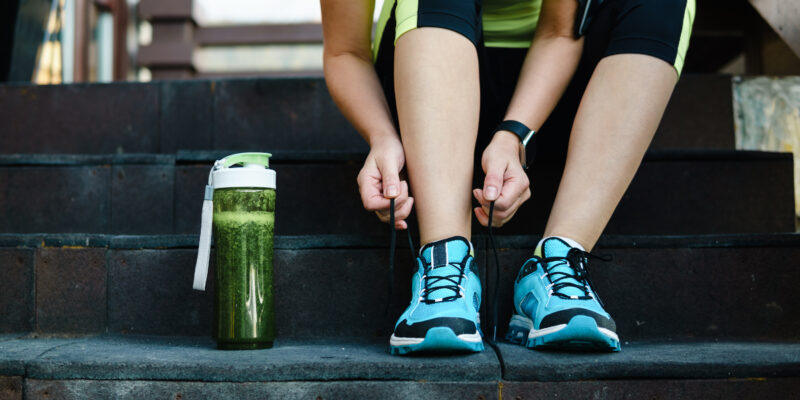 Green detox smoothie cup and woman lacing running shoes before workout